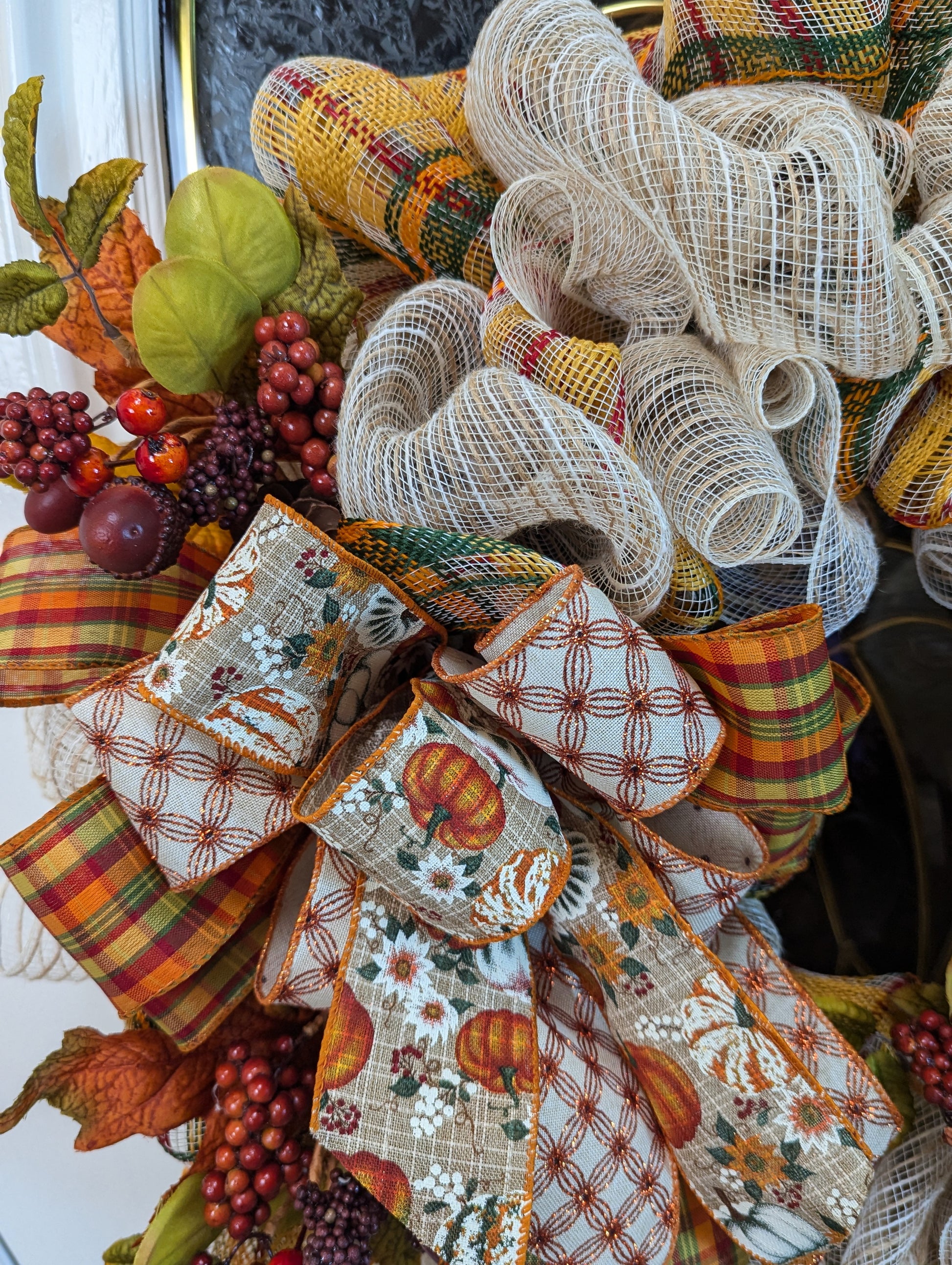 Fall Wreath with beige deco mesh base with a curly Q design. There are 2 matching bows with orange, gold, green and beige patterned ribbons. It has a velveteen pumpkin, acorns, pinecones and fall foliage.
