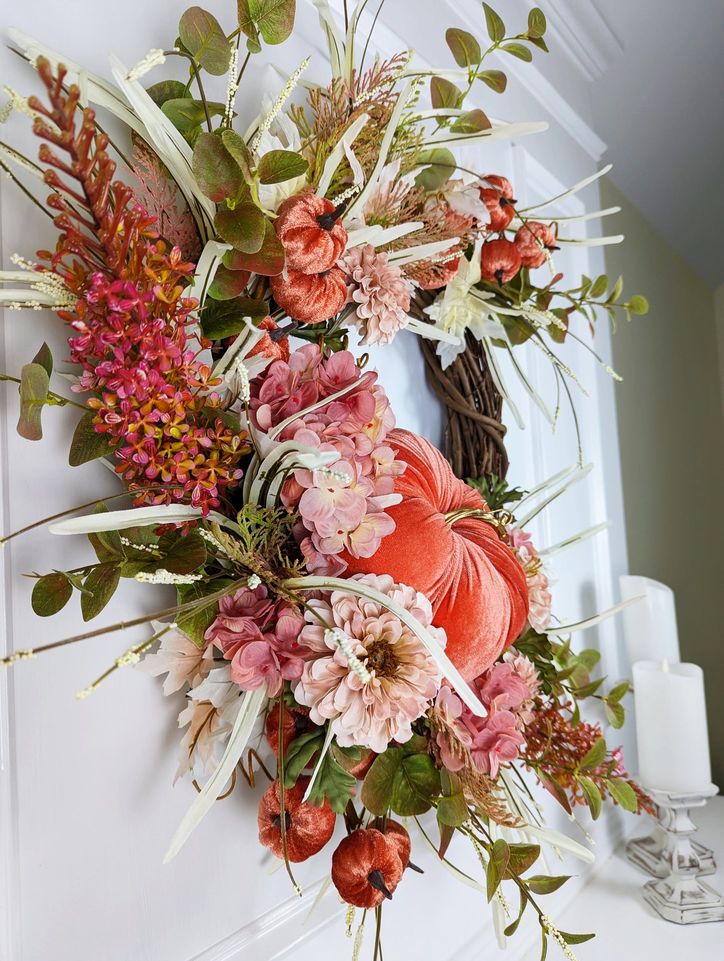 Fall wreath with a pretty orange colored velveteen pumpkin surrounded by soft pink and flesh toned hydrangeas and chrysanthemums. A brown grapevine base with wispy light green and ivory grass and maple leaves.