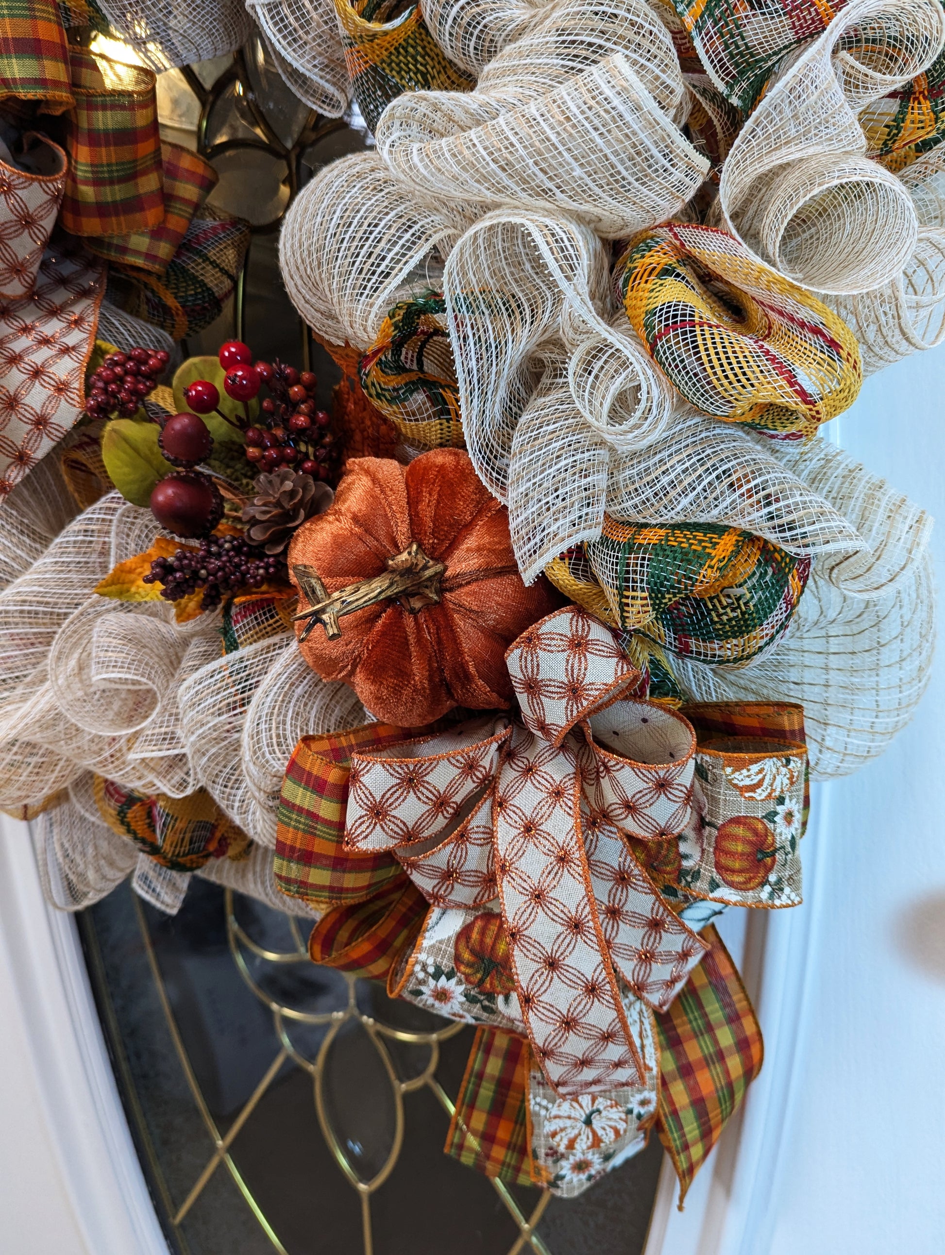 Fall Wreath with beige deco mesh base with a curly Q design. There are 2 matching bows with orange, gold, green and beige patterned ribbons. It has a velveteen pumpkin, acorns, pinecones and fall foliage.