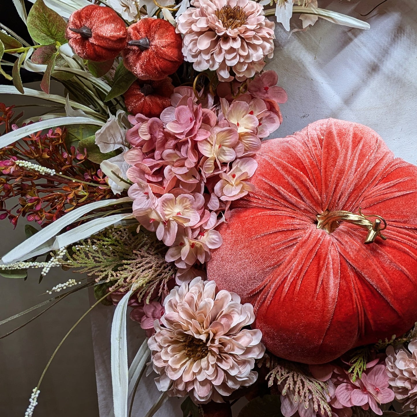 Fall wreath with a pretty orange colored velveteen pumpkin surrounded by soft pink and flesh toned hydrangeas and chrysanthemums. A brown grapevine base with wispy light green and ivory grass and maple leaves.