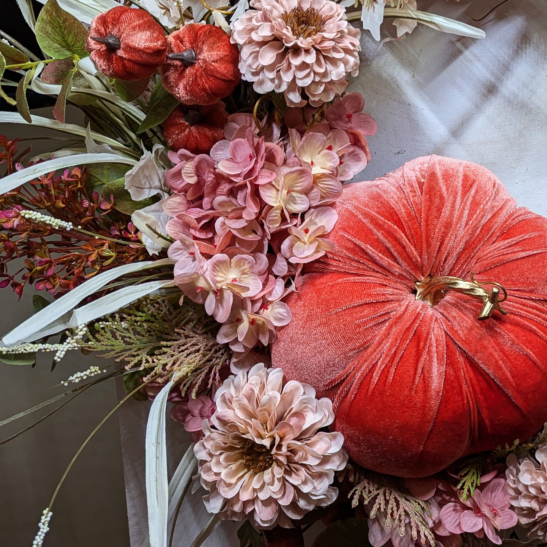 Fall wreath with a pretty orange colored velveteen pumpkin surrounded by soft pink and flesh toned hydrangeas and chrysanthemums. A brown grapevine base with wispy light green and ivory grass and maple leaves.