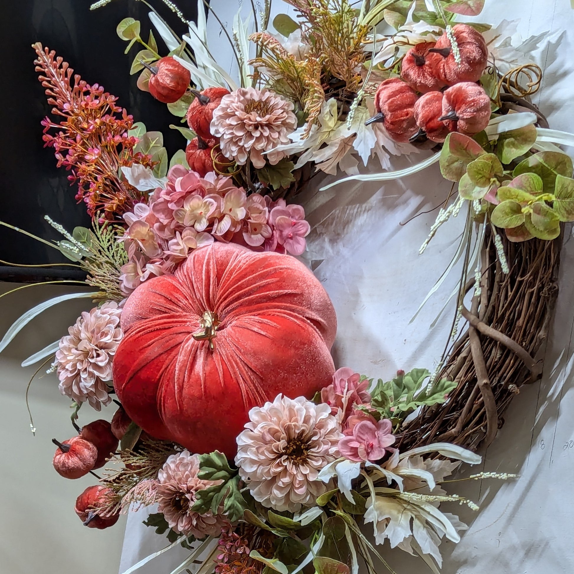 Fall wreath with a pretty orange colored velveteen pumpkin surrounded by soft pink and flesh toned hydrangeas and chrysanthemums. A brown grapevine base with wispy light green and ivory grass and maple leaves.
