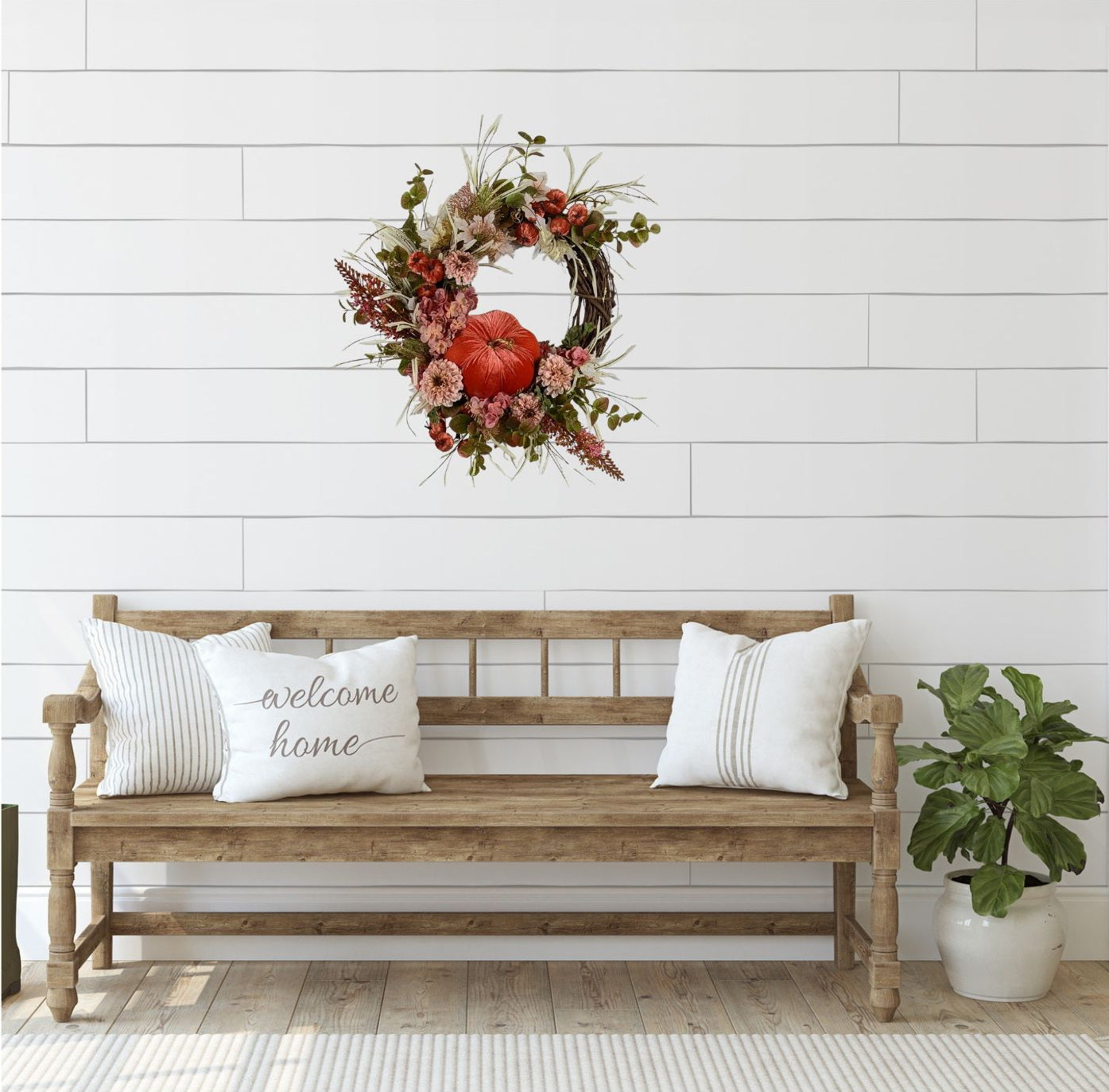 Fall wreath with a pretty orange colored velveteen pumpkin surrounded by soft pink and flesh toned hydrangeas and chrysanthemums. A brown grapevine base with wispy light green and ivory grass and maple leaves. Styled in a foyer vignette. 