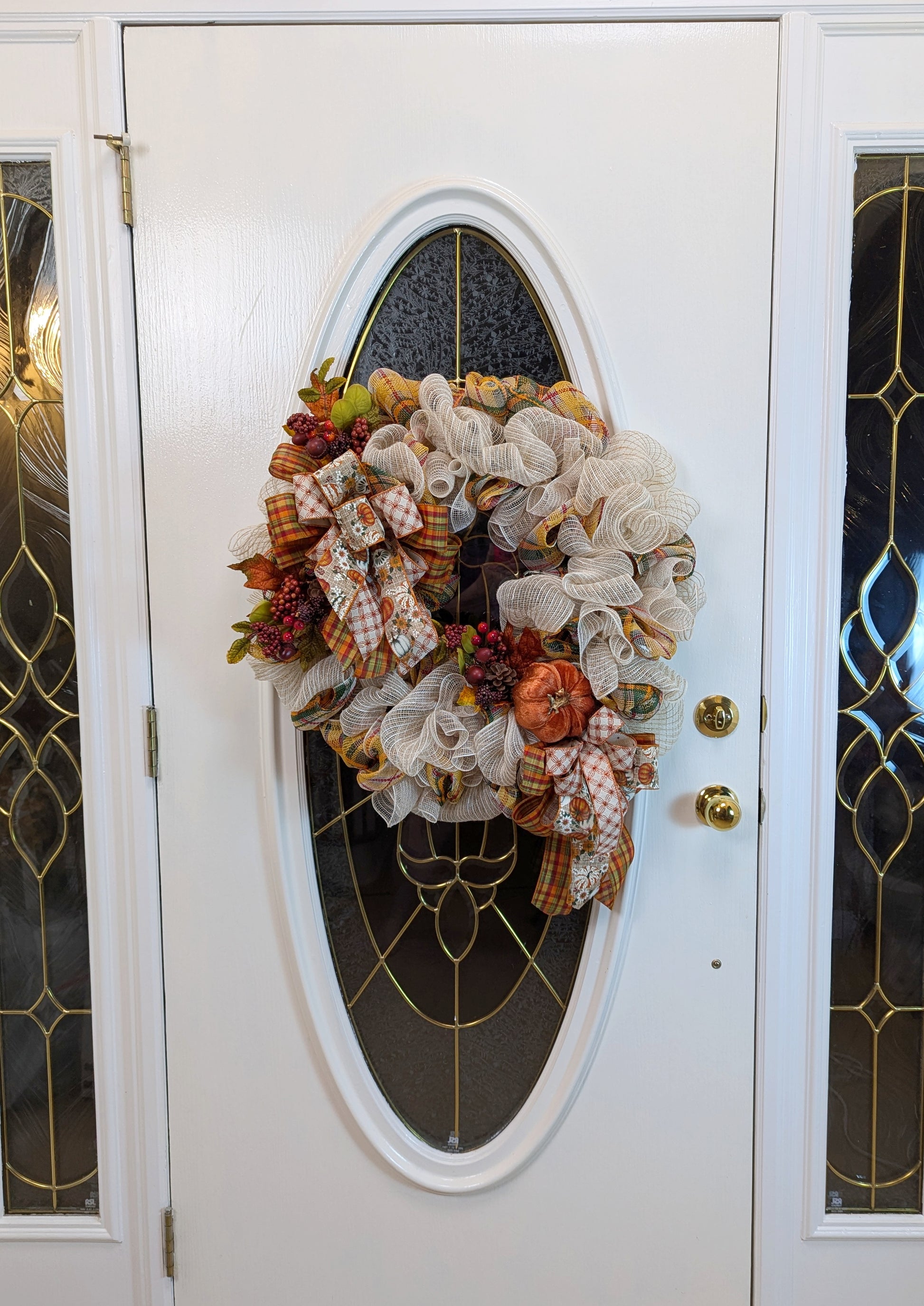 Fall Wreath with beige deco mesh base with a curly Q design. There are 2 matching bows with orange, gold, green and beige patterned ribbons. It has a velveteen pumpkin, acorns, pinecones and fall foliage.