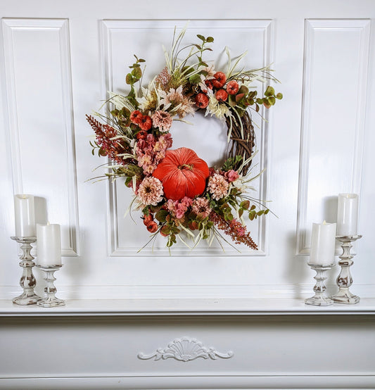 Fall wreath with a pretty orange colored velveteen pumpkin surrounded by soft pink and flesh toned hydrangeas and chrysanthemums. A brown grapevine base with wispy light green and ivory grass and maple leaves.